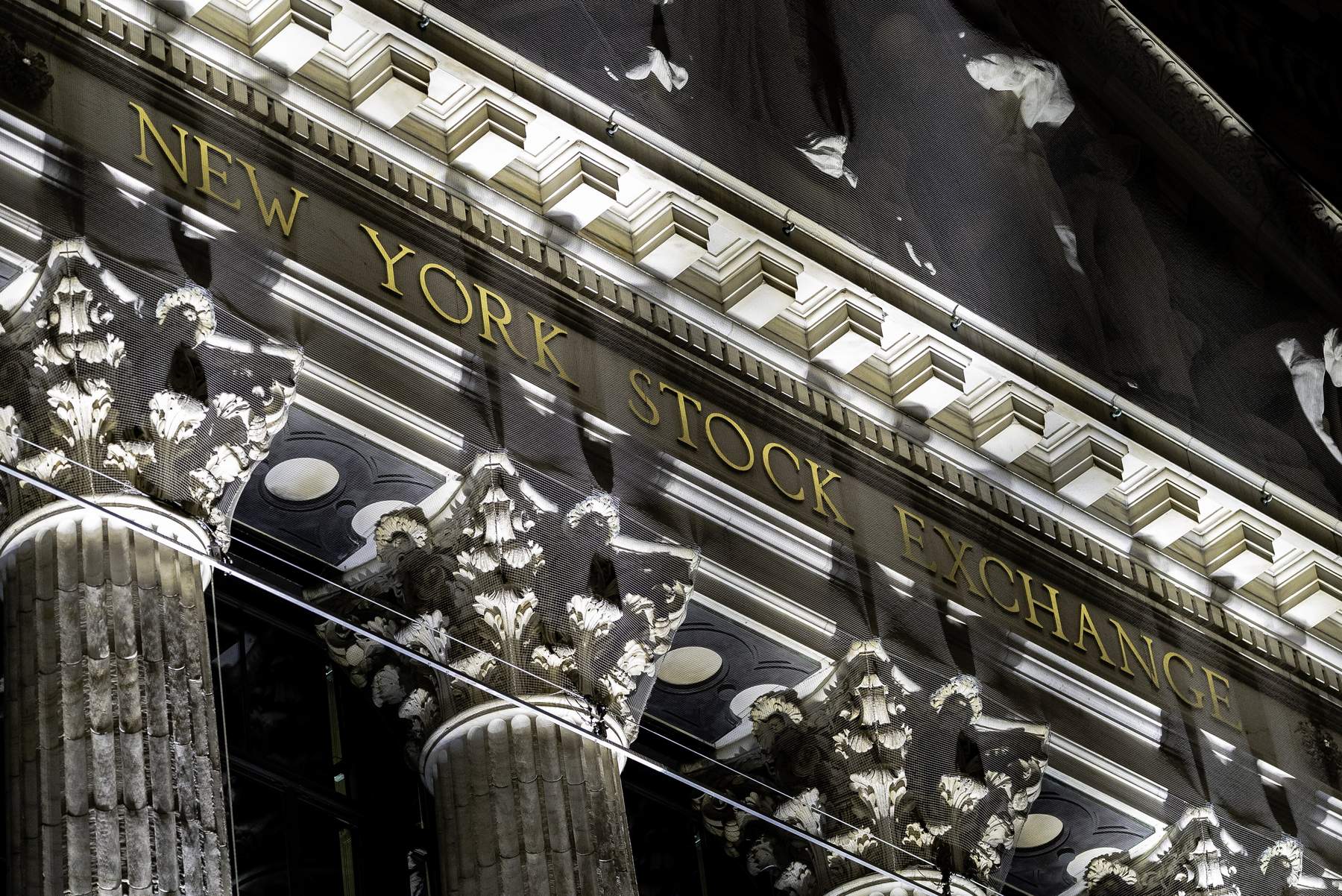 Gilded “NEW YORK STOCK EXCHANGE” lettering and the tops of Corinthian columns on the exchange’s facade, lit at night and partly behind mesh netting