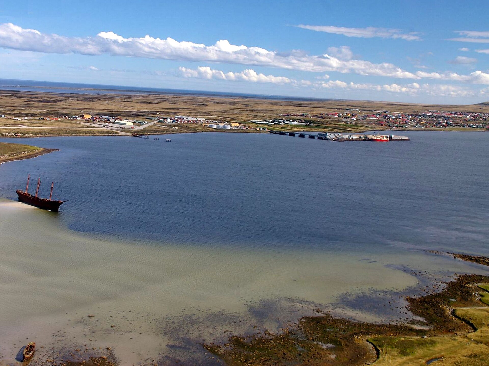 Illustrative aerial view across Stanley Harbour in the Falkland Islands, with the town of Stanley on the far shore and the wreck of a sailing ship in the shallows