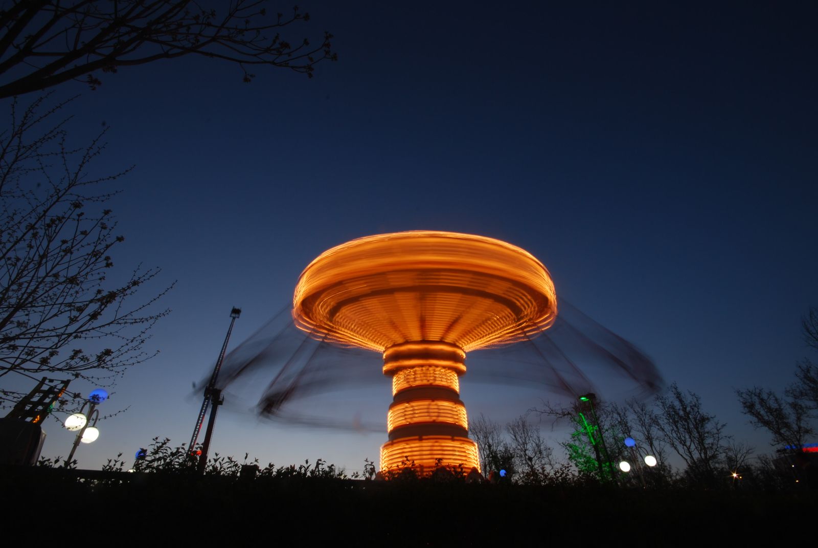A spinning swing ride glowing orange against a deep-blue dusk sky, blurred by a long exposure, with bare trees in silhouette — illustrative stock