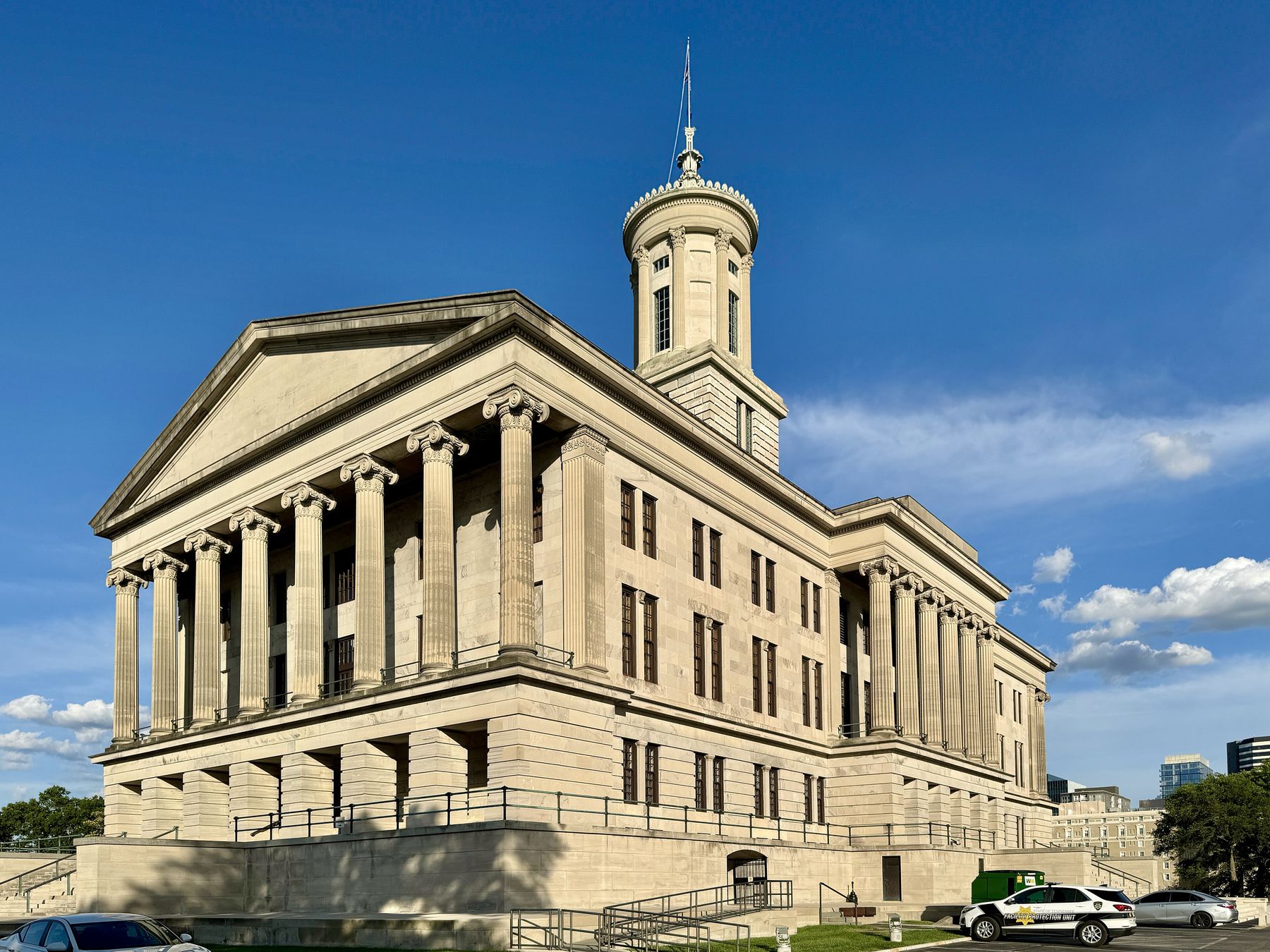 Tennessee State Capitol, a pale stone Greek Revival building with a columned portico and a cupola, under a blue sky, with cars parked along the street at right