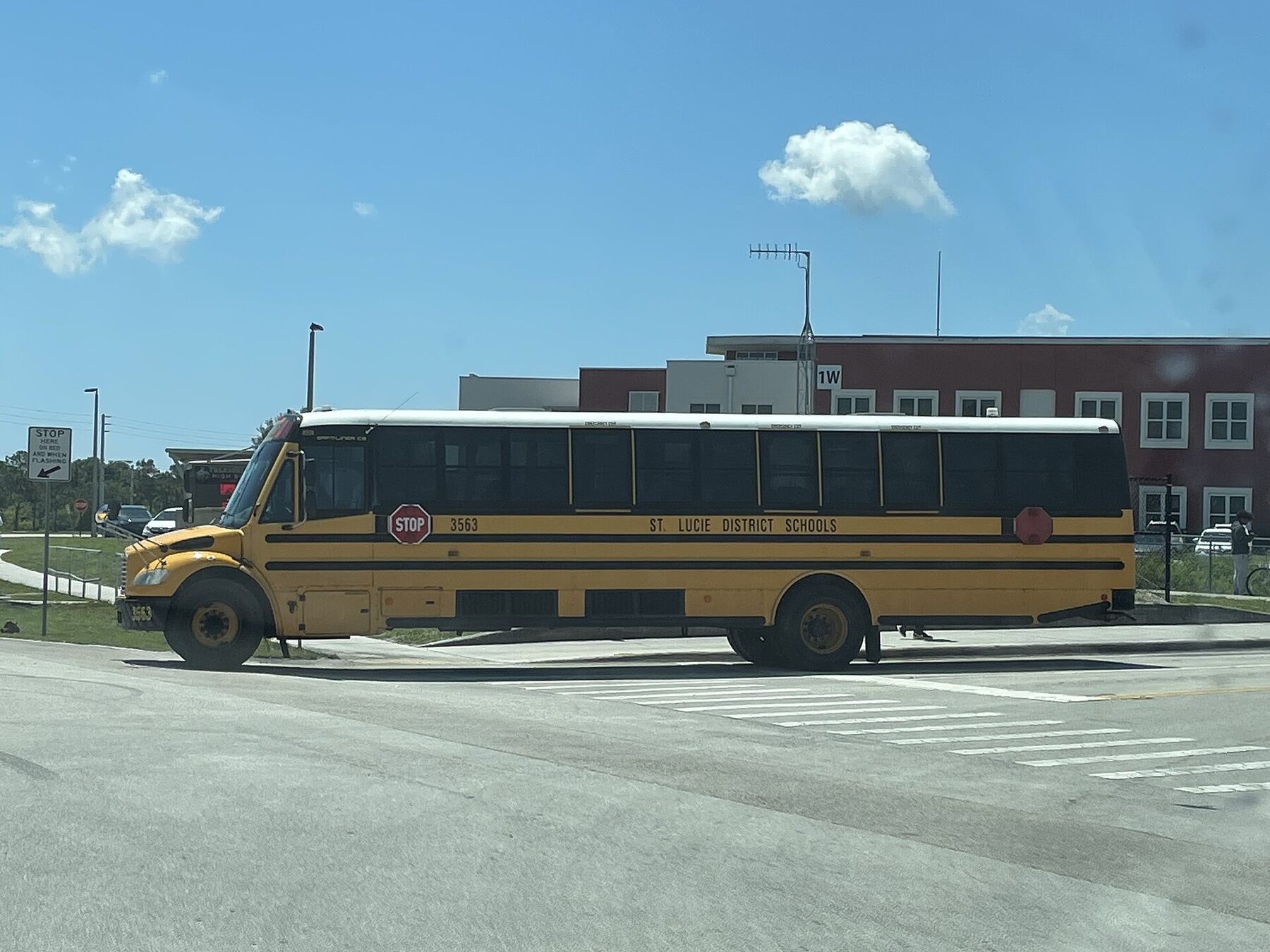 Yellow school bus marked “St. Lucie District Schools” and numbered 3563, stopped near a crosswalk in front of a red-brick school building under a blue sky