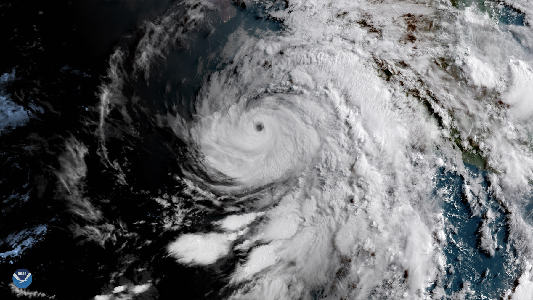 Satellite image of a large hurricane with a small, clearly defined eye over the dark ocean, with cloud bands spreading toward land at right and a NOAA logo at lower left