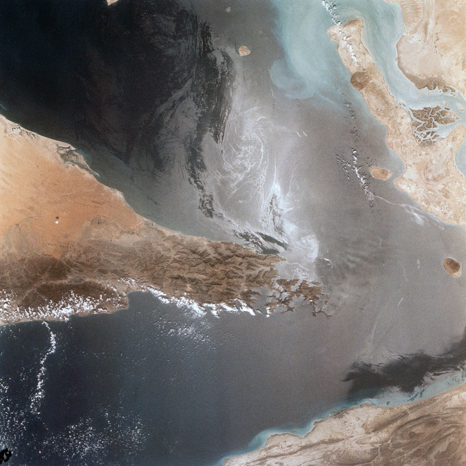 Photograph of the Strait of Hormuz taken from Space Shuttle Columbia in 1982, with the mountainous tip of Oman's Musandam Peninsula reaching in from the left, Iran's coast and Qeshm Island at right, and sunglint on the water between them — illustrative