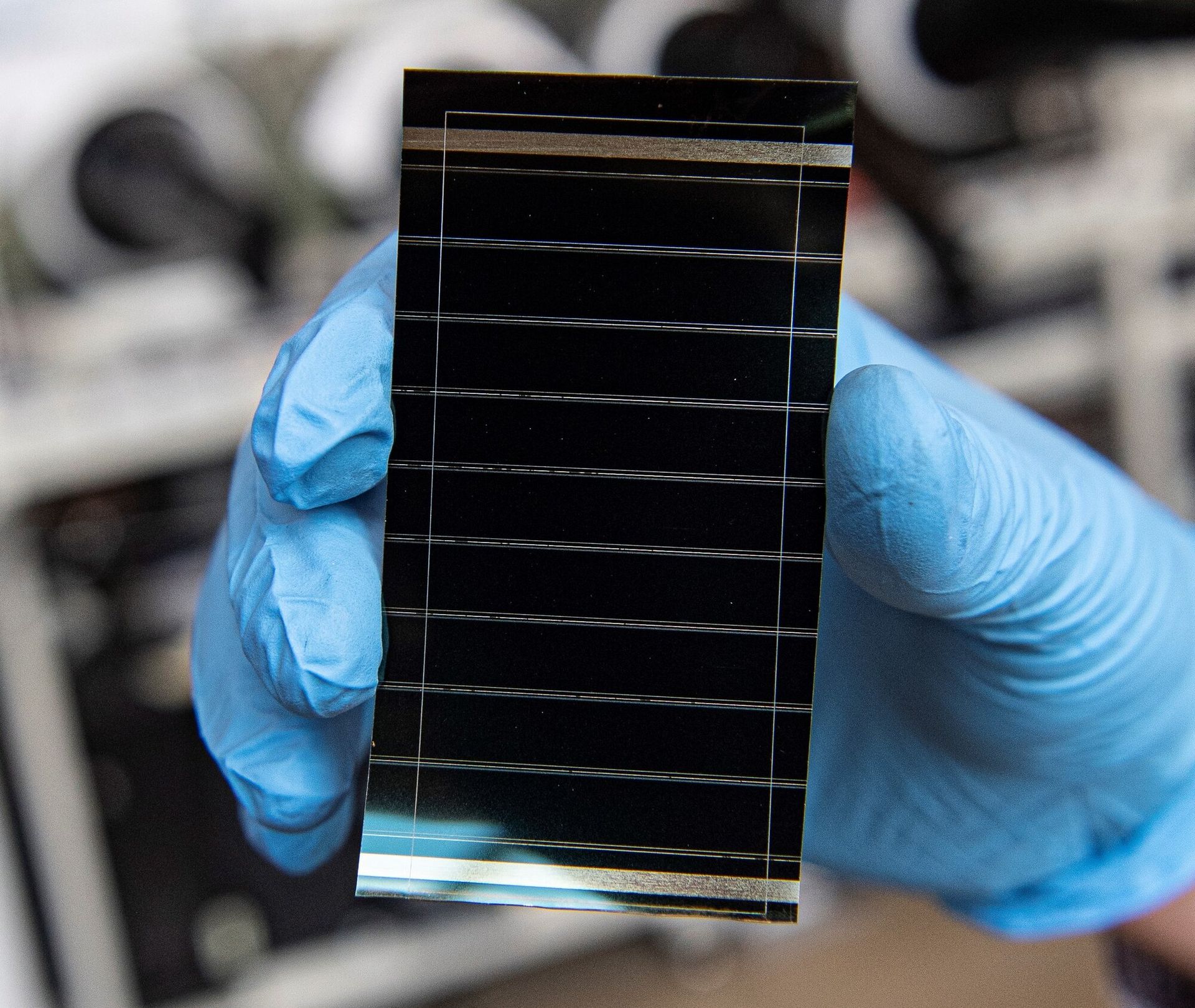 A gloved hand holds a small, dark rectangular perovskite solar cell crossed by thin horizontal lines in a laboratory — illustrative, not the Nanjing tandem device