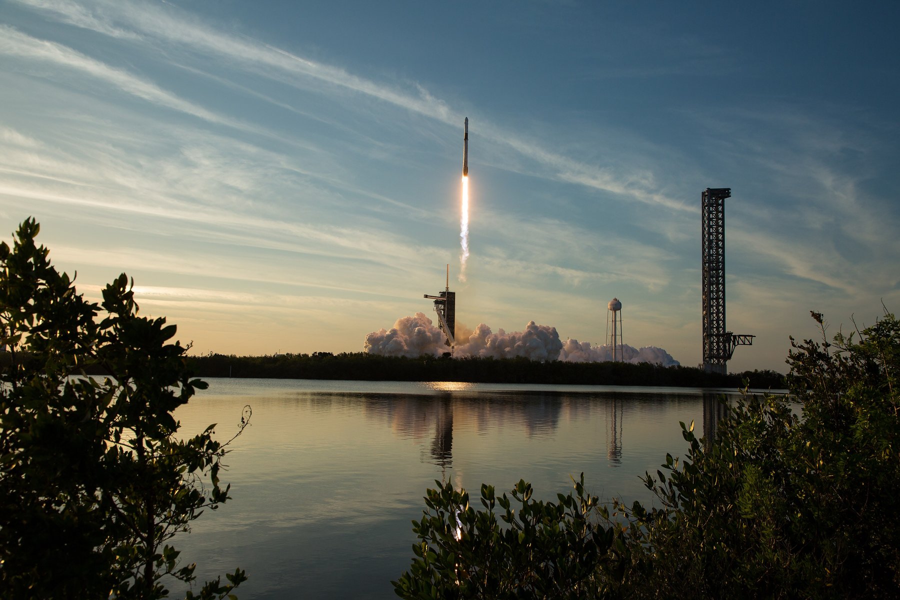 A Falcon 9 rocket rising on a bright exhaust plume above a launch pad and a billowing steam cloud, with the scene reflected in water and shrubs in the foreground