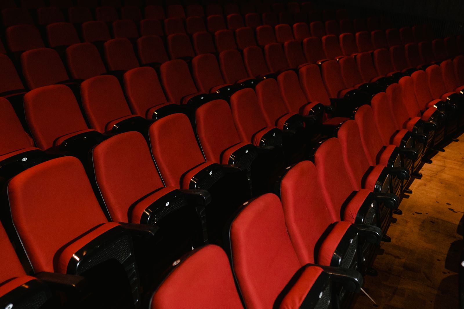 Rows of empty red upholstered auditorium seats with black armrests curving away into darkness beside a wooden floor — illustrative stock
