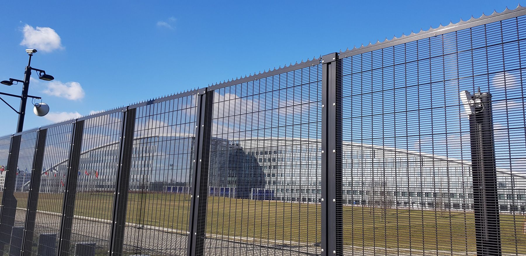 NATO headquarters building in Brussels seen through a tall black security fence with a spiked top, with a lawn beyond the fence and a lamp post at left, under a blue sky