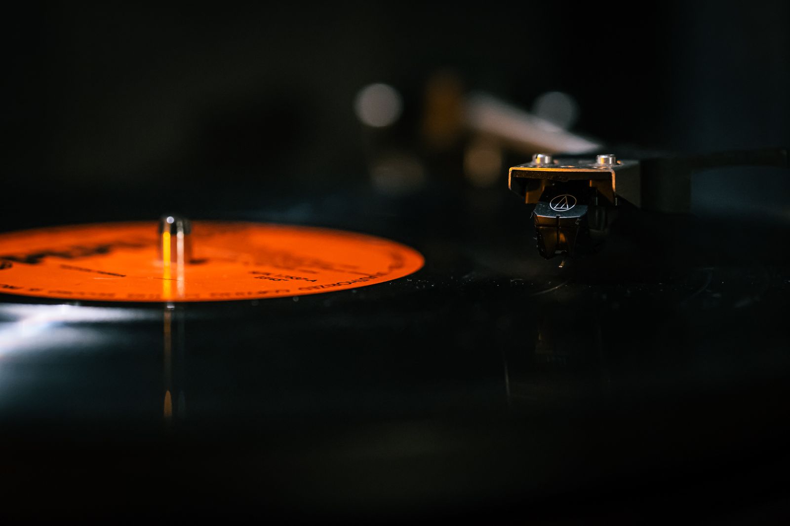 Close-up of a turntable cartridge and stylus resting on a spinning black vinyl record with an orange center label, against a dark background — illustrative stock
