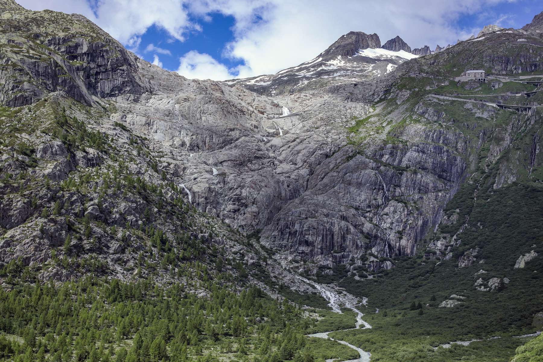 Bare, ice-scoured grey rock and a meltwater stream in a valley below a mountain ridge, with patches of snow near the peaks and a small building on the ridge