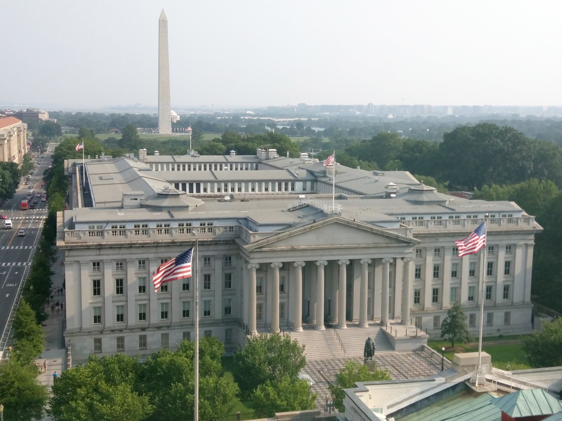Elevated view of the U.S. Treasury Department building with American flags flying in front and the Washington Monument in the hazy distance