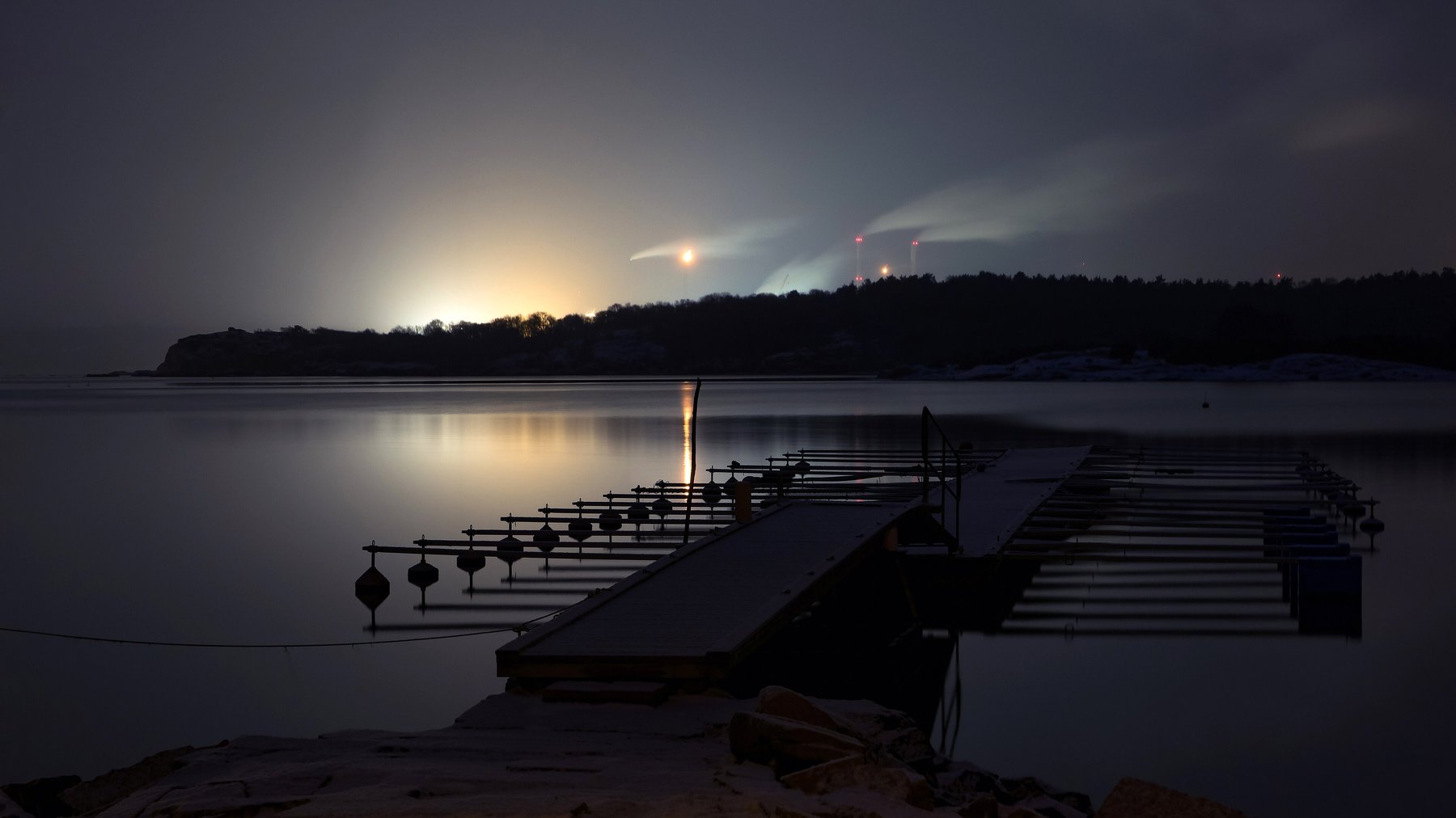 Night view across a calm fjord with a floating dock in the foreground and the lights and flare stacks of a distant oil refinery on the far shore